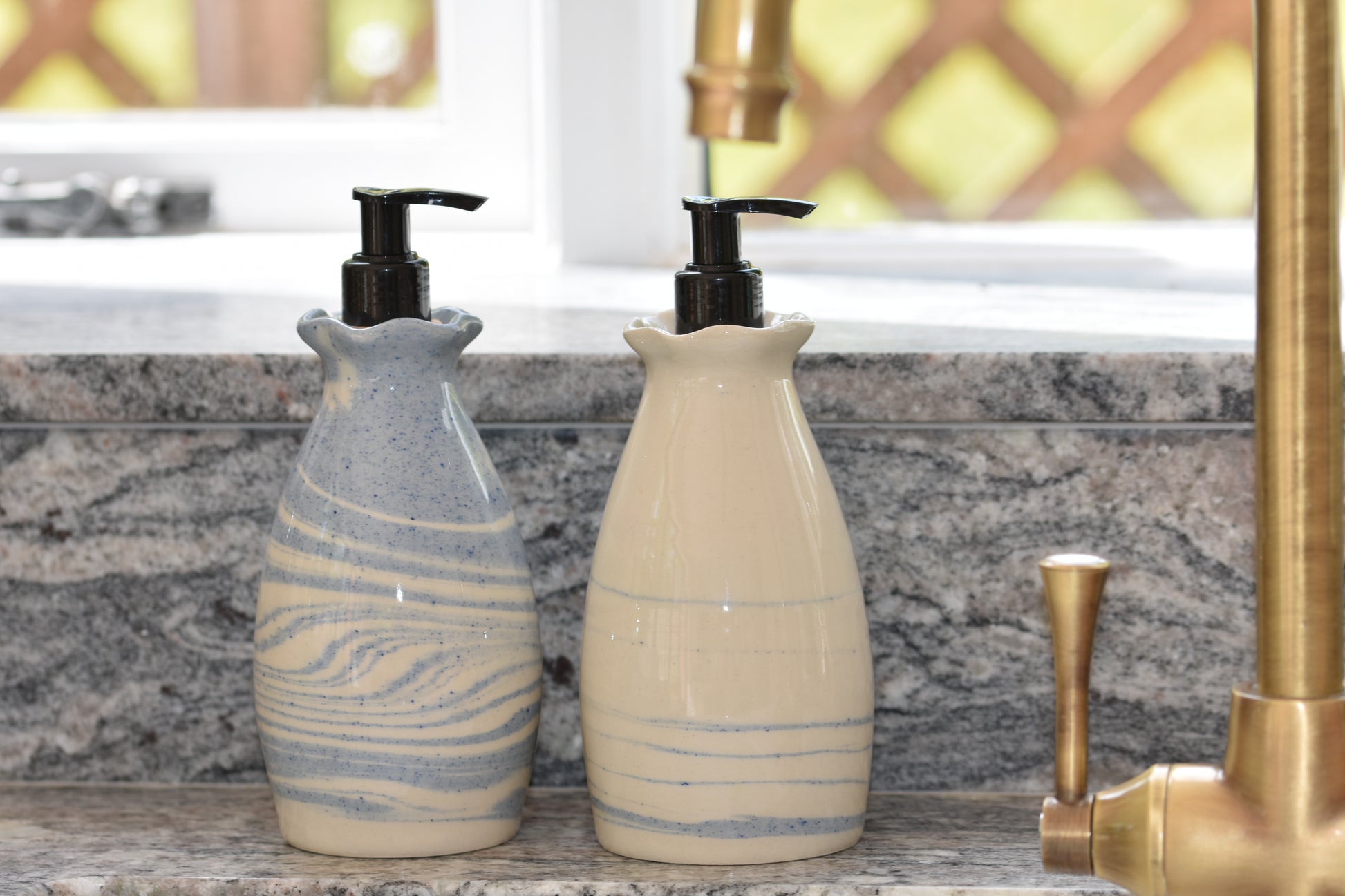 Two Ceramics by Sunny soap dispensers in white and speckled sky blue stoneware with black pumps at the sink with brass tap and granite worktop.