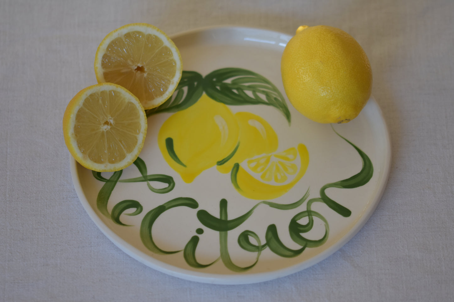 Le Crabe Le Citron ceramic plate with painted lemon and French writing, shown with lemons on a beige background.