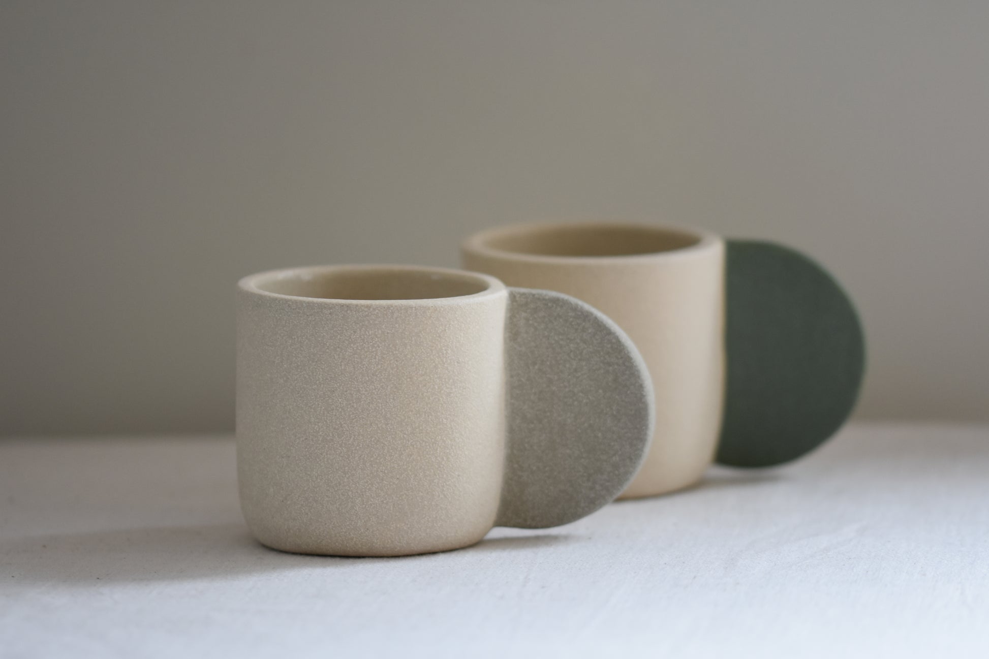 Brutes ceramics stoneware medium signature mug in sand with grey handle and textured finish, in front of Brutes ceramics stoneware medium signature mug with granite green handle.
