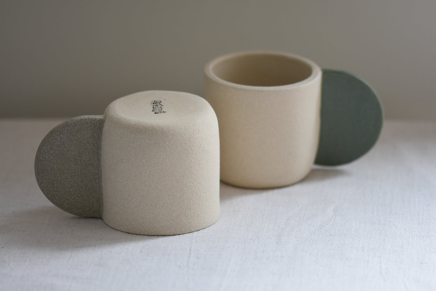 Brutes ceramics stoneware medium signature mug in sand with grey handle and textured finish, in front of Brutes ceramics stoneware medium signature mug with granite green handle.