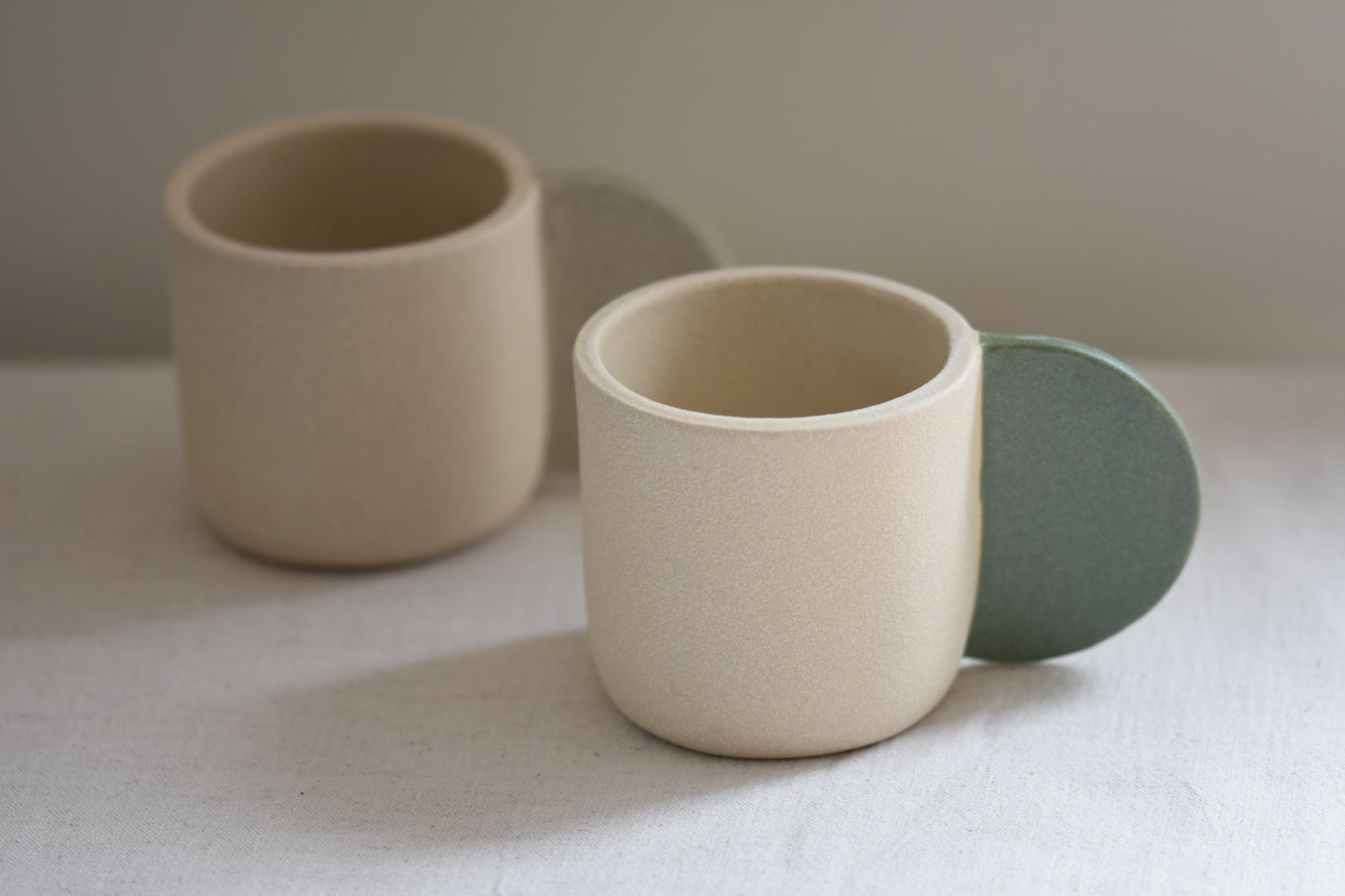 Brutes ceramics stoneware medium signature mug in textured finish with granite green glazed handle, in front of Brutes ceramics stoneware medium signature mug in sand.