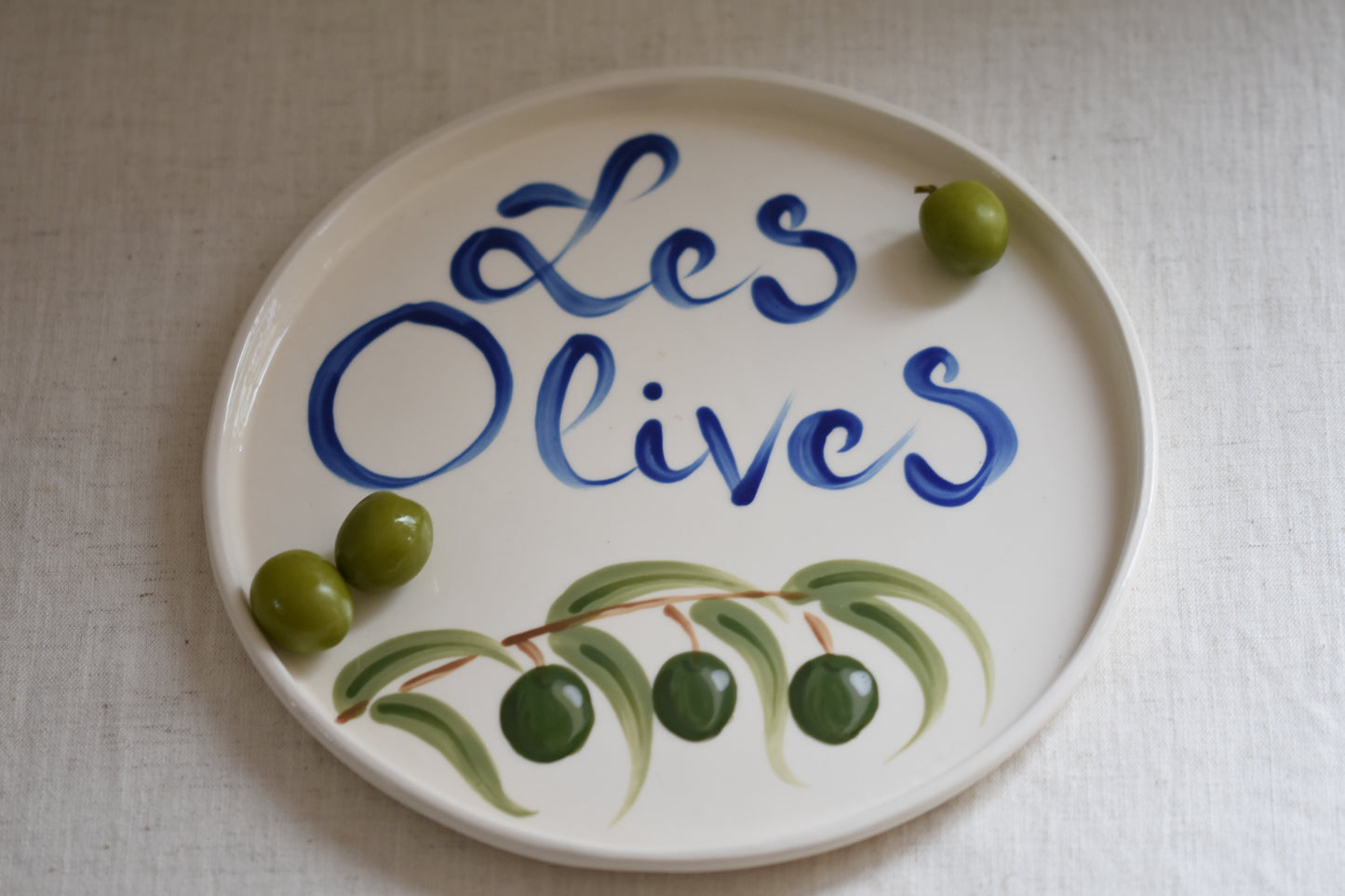Le Crabe Les Olives ceramic plate with painted olives and French writing, shown with green olives on the plate, on a beige background.