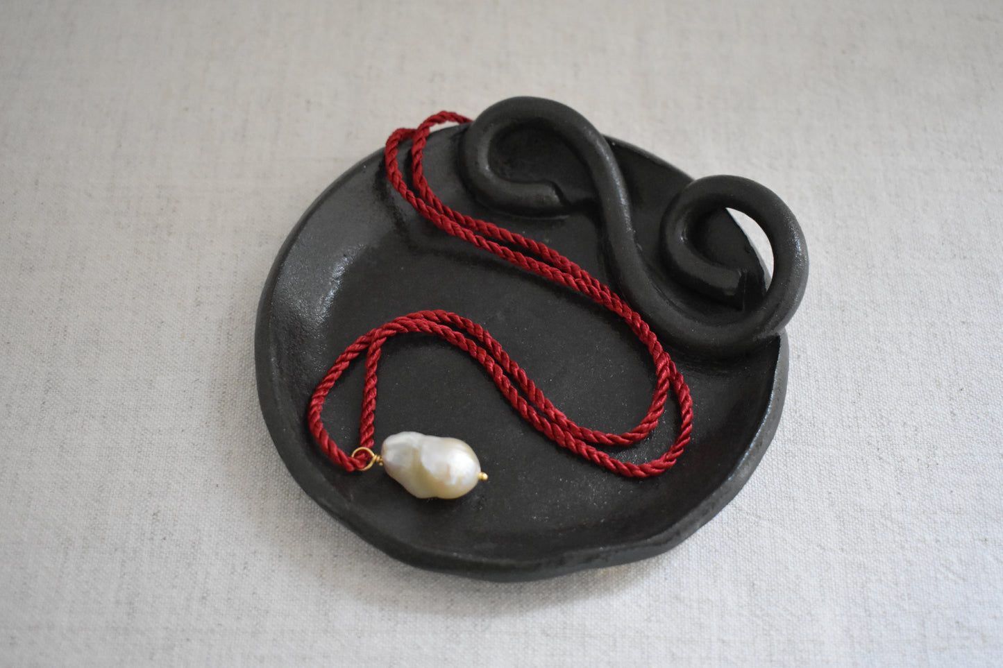 Sandralexandra baroque pearl on dark red silk twisted cord necklace shown on We Are Clay black ceramic spiral Wee Dish, with a beige background.