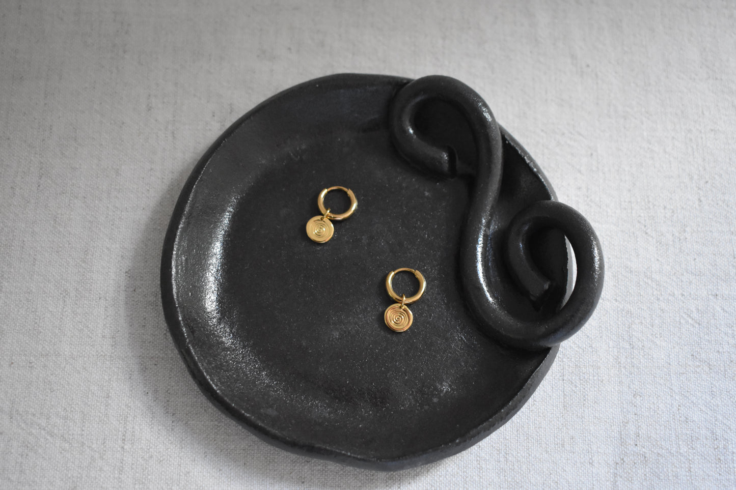Sandralexandra Mini Sea Urchin spiral hoop earrings in gold on We Are Clay black ceramic spiral Wee Dish with a beige background.