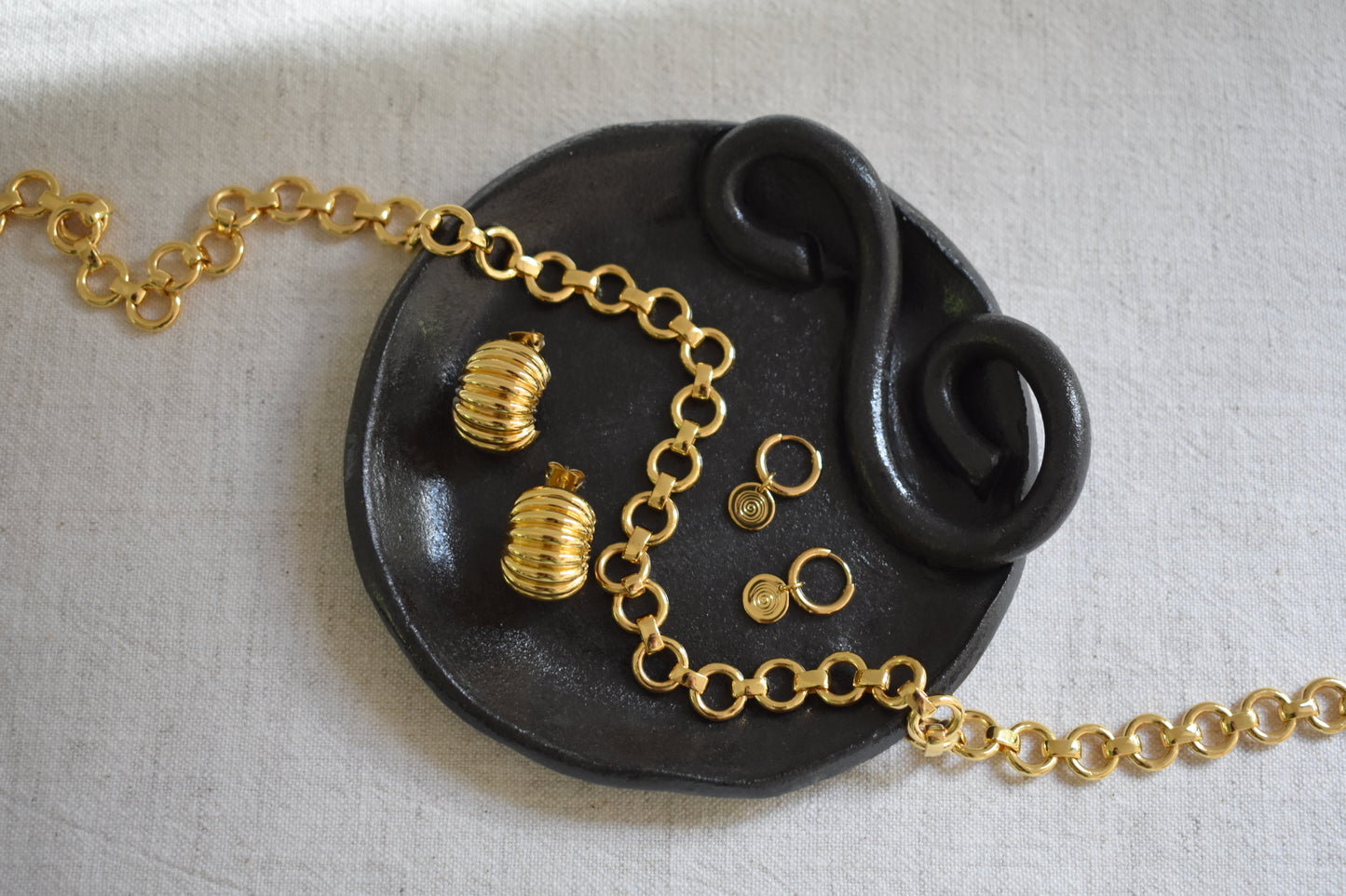 We Are Clay Wee Dish in black ceramic with spiral design holding Daphine Baby Bea necklace, Daphine Clara hoops and Sandralexandra Mini Sea Urchin hoops in gold.