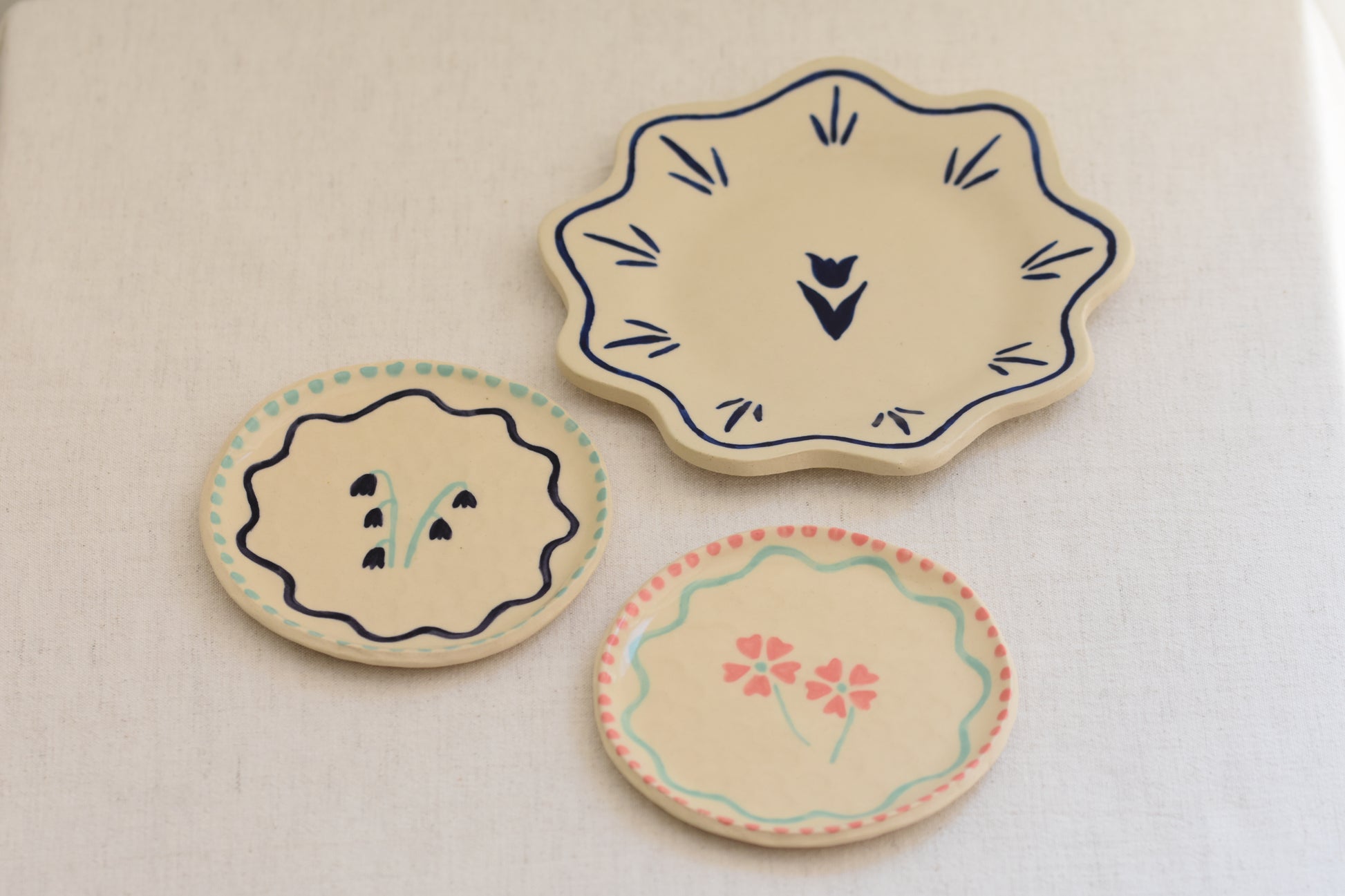Blume Ceramics white stoneware small plates with hand painted floral design in pink and green, and bluebell design in navy and green, alongside a white stoneware petal shaped plate with hand painted tulip design in navy.