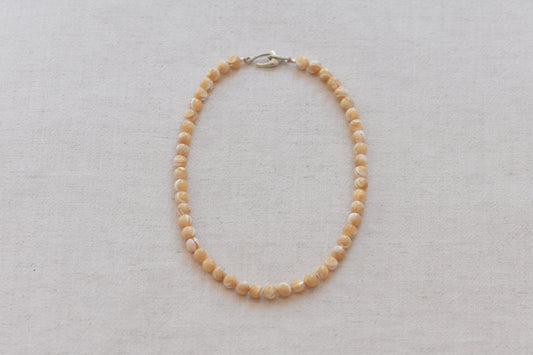 By my hands Sandy Bei Necklace of opalescent light yellow Mother of Pearl beads, with a hand-carved sterling silver fish hook closure, on a beige background.