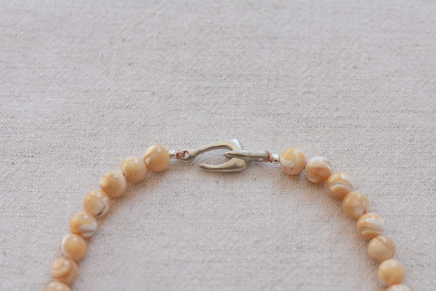 By my hands Sandy Bei Necklace of opalescent light yellow Mother of Pearl beads, with a hand-carved sterling silver fish hook closure, close up, on a beige background.