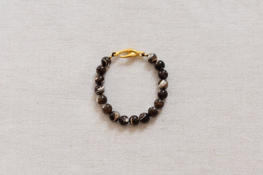 By my hands Burnt Umber Bei Bracelet of opalescent dark brown Mother of Pearl beads, with a hand-carved gold vermeil fish hook closure, on a beige background.