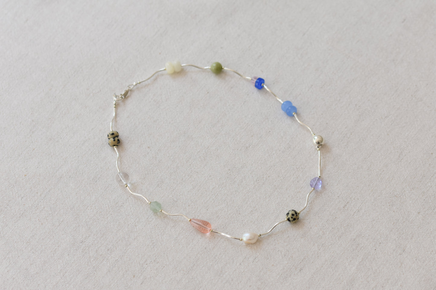 By my hands Fluorescence Ripple Necklace with wavy sterling silver ripples and gemstone beads including peridot, dalmatian jasper, freshwater pearl, and coloured jade, on a beige background.