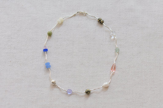 By my hands Fluorescence Ripple Necklace with wavy sterling silver ripples and gemstone beads including peridot, dalmatian jasper, freshwater pearl, and coloured jade, on a beige background.