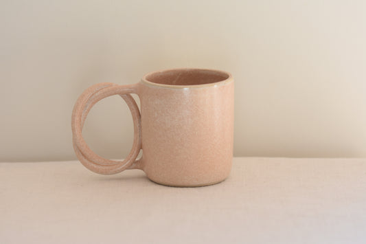We Are Clay blush pink ceramic mug with pretzel shaped handle on a beige background.