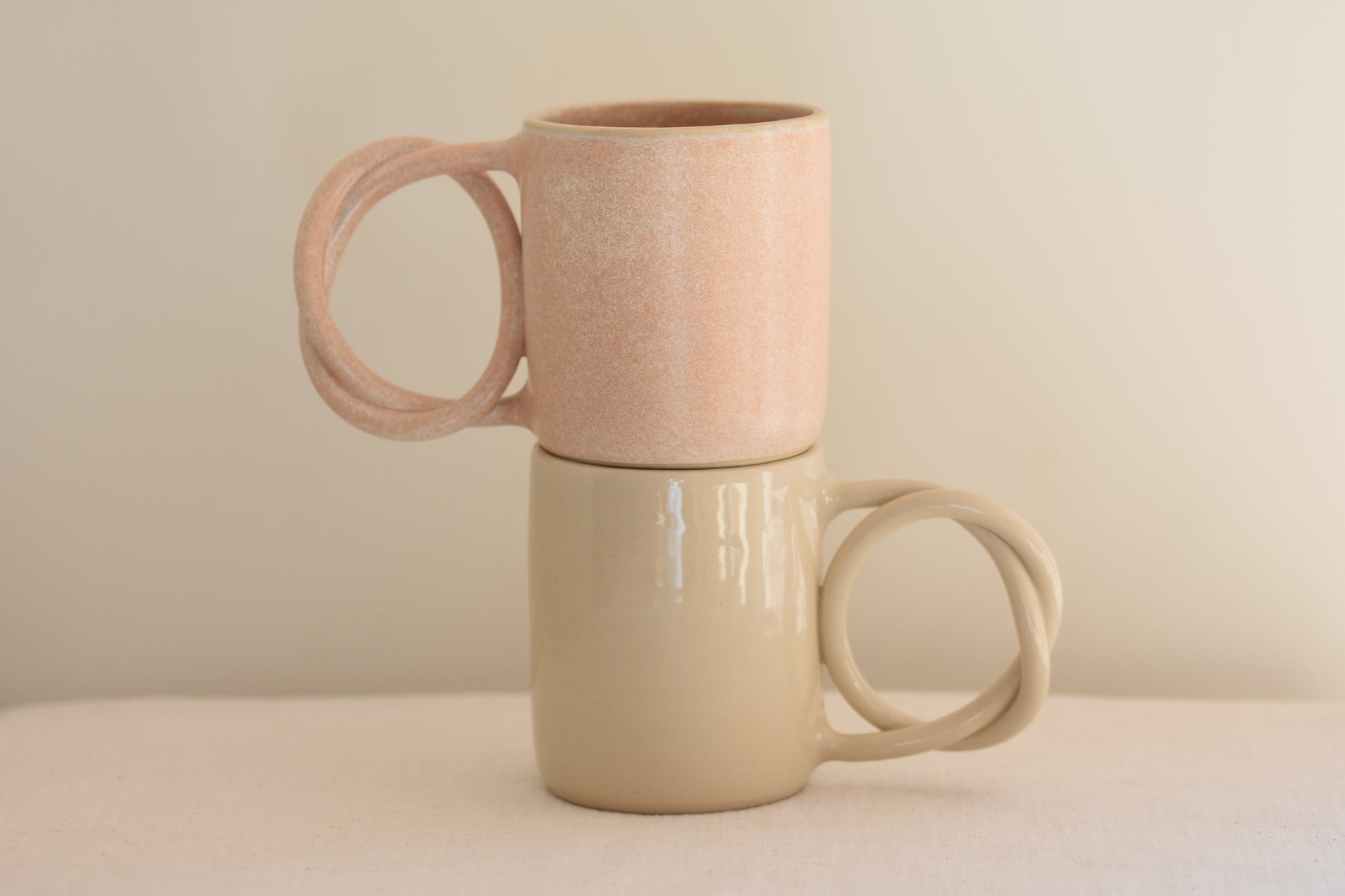 We Are Clay blush pink ceramic mug with pretzel shaped handle, stacked on top of white ceramic mug with pretzel shaped handle, on a beige background.