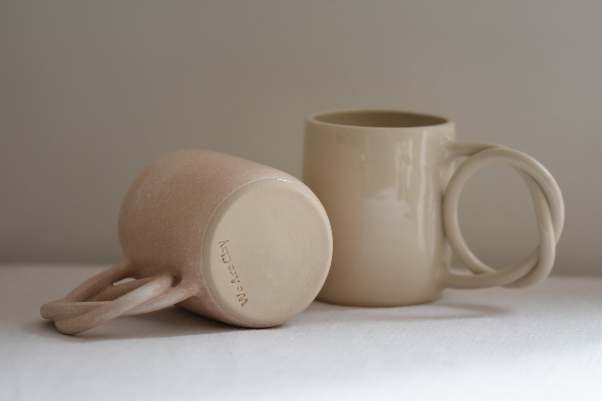 We Are Clay blush pink ceramic mug with pretzel shaped handle, lying down to show makers mark, next to white ceramic mug with pretzel shaped handle, on a beige background.