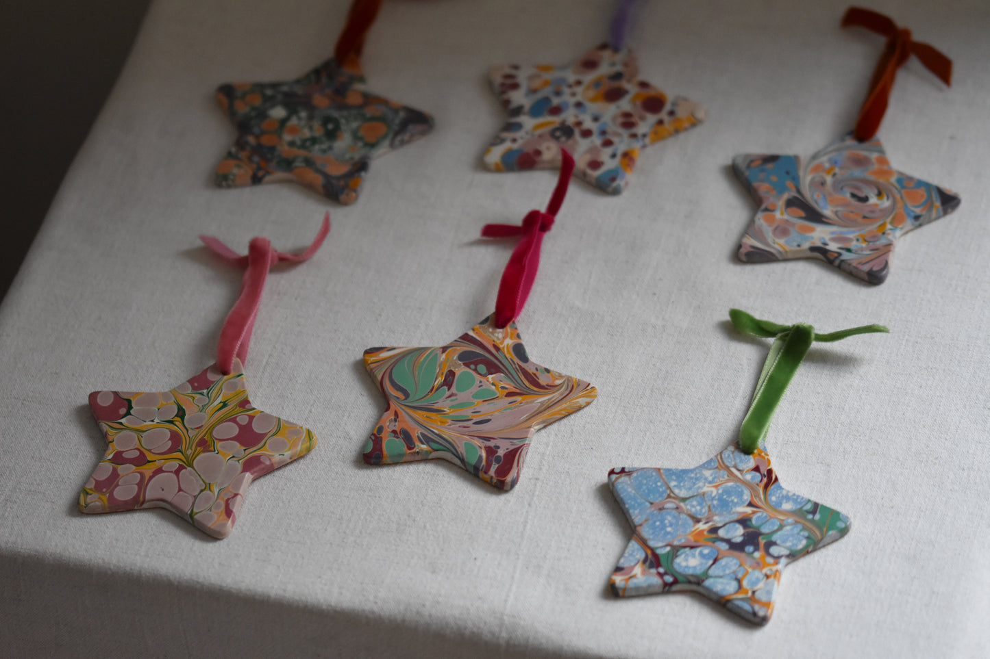 Six Jemma Lewis marbled ceramic star decorations with colourful velvet ribbons on a beige background.