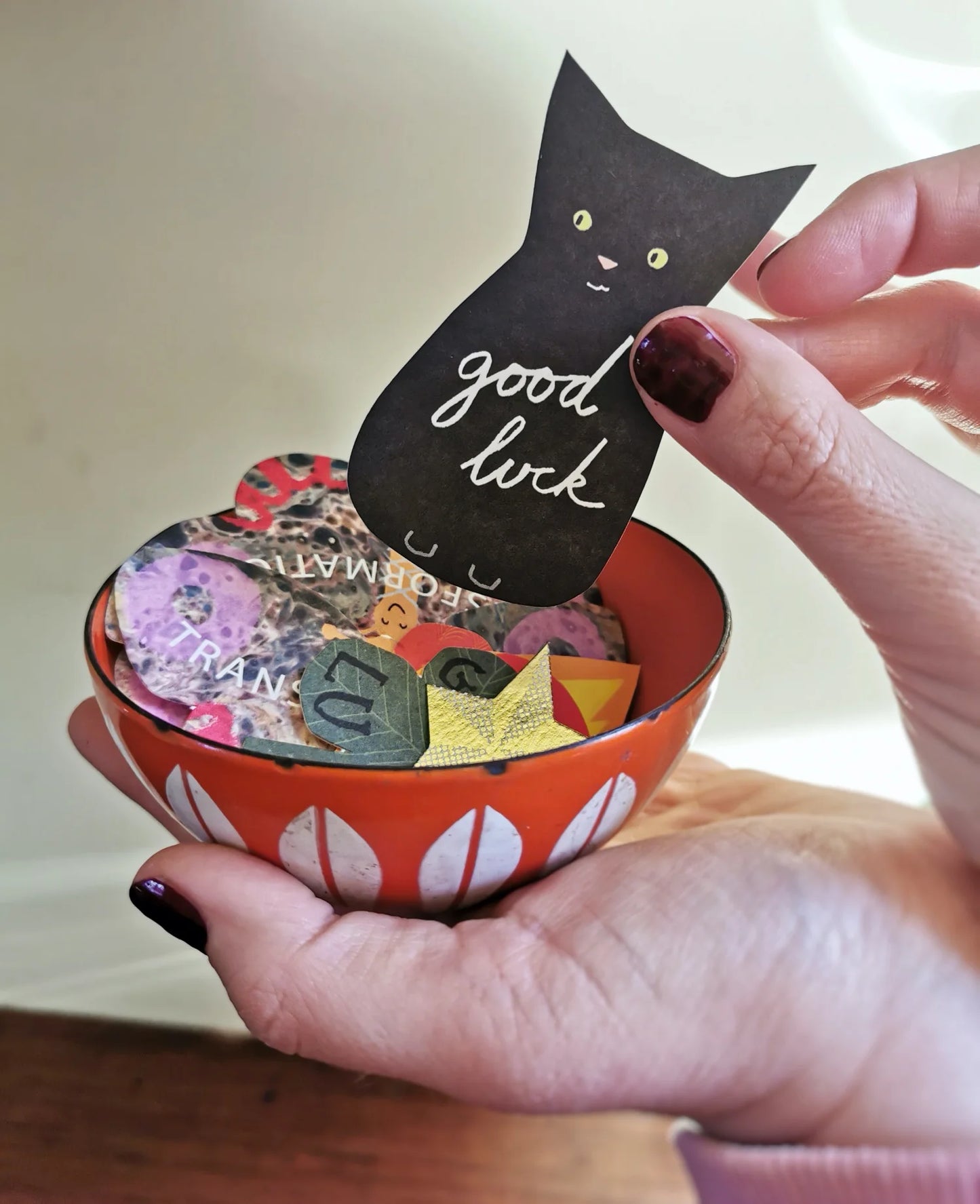 Hands holding a bowl of Hadley Paper Goods Manifestation Charms, multicolour card shapes including a black good luck cat, with a cream background.