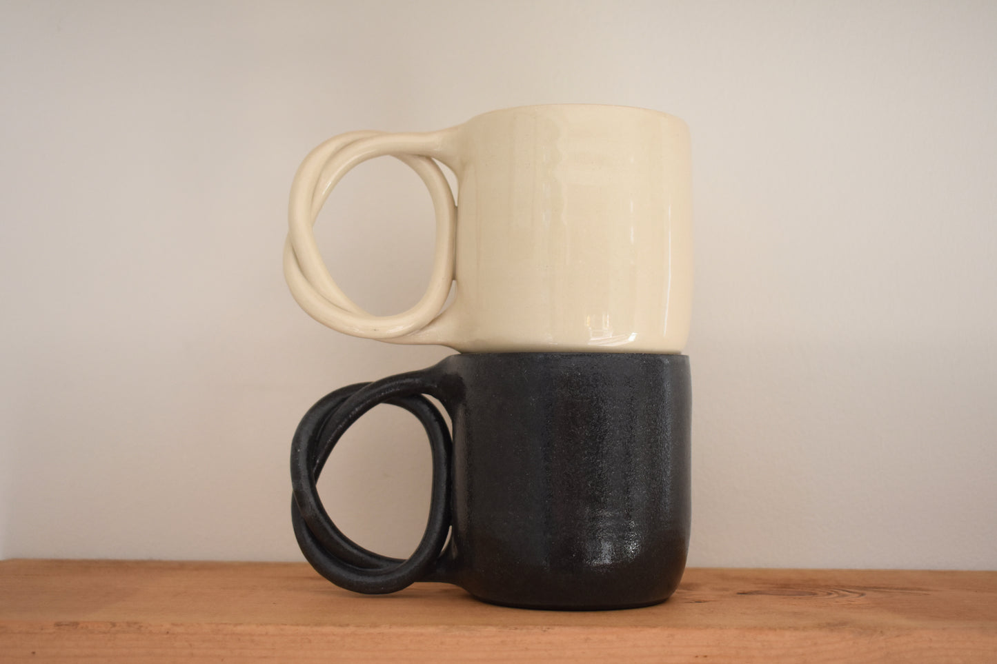 We Are Clay ceramic Pretzel Mugs with twisted pretzel handles in black and white, stacked.