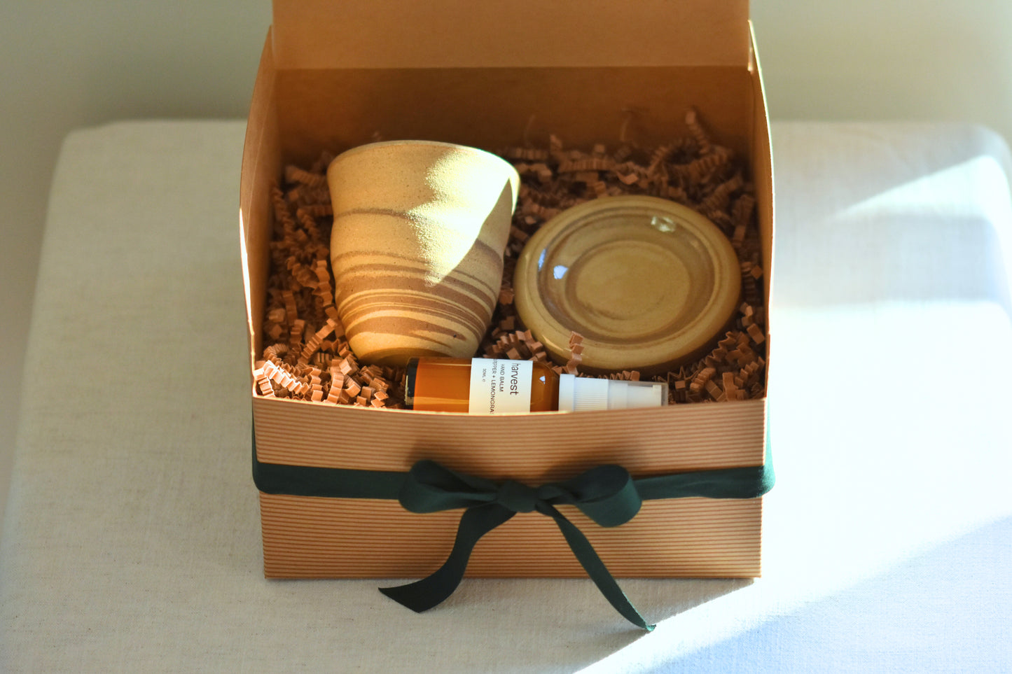 RORA kraft gift box with green ribbon containing a Ceramics by Sunny spiral cup, doughnut plate and Harvest hand balm, on a beige background.