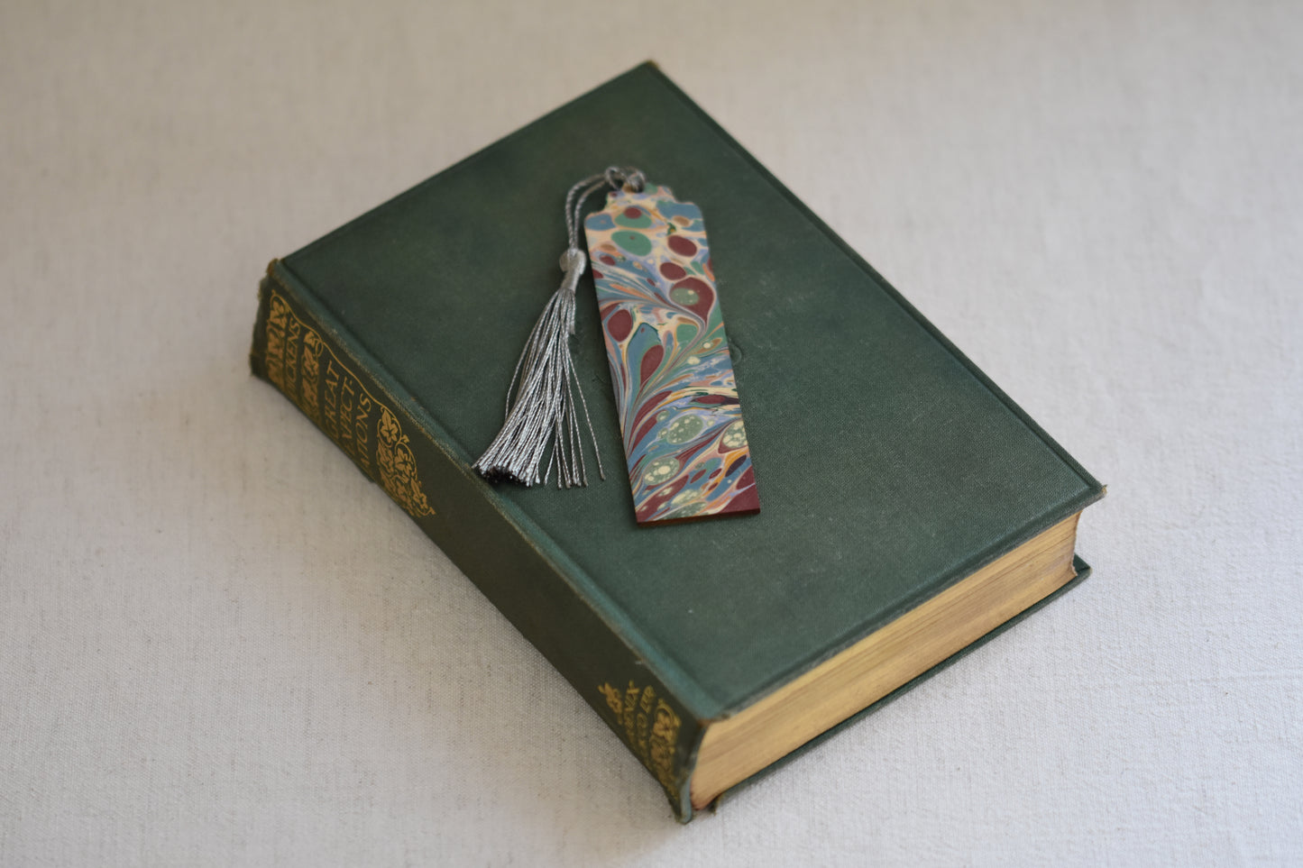 Jemma Lewis colourful marbled wooden bookmark with grey tassel on a vintage green and gold book, with a beige background.