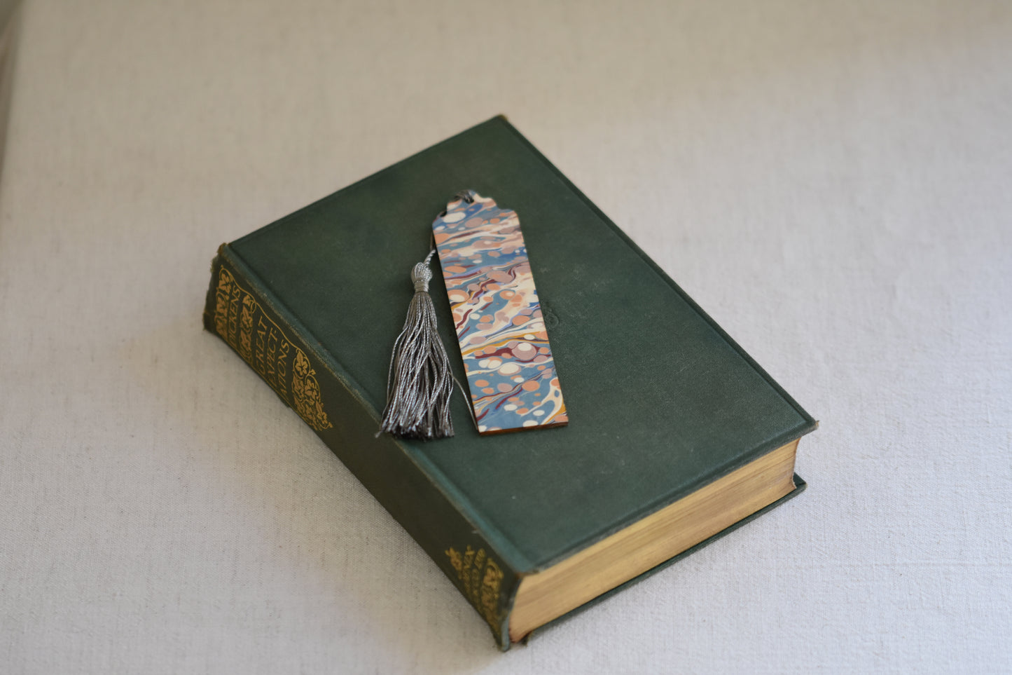 Jemma Lewis colourful marbled wooden bookmark with grey tassel on a vintage green and gold book, with a beige background.
