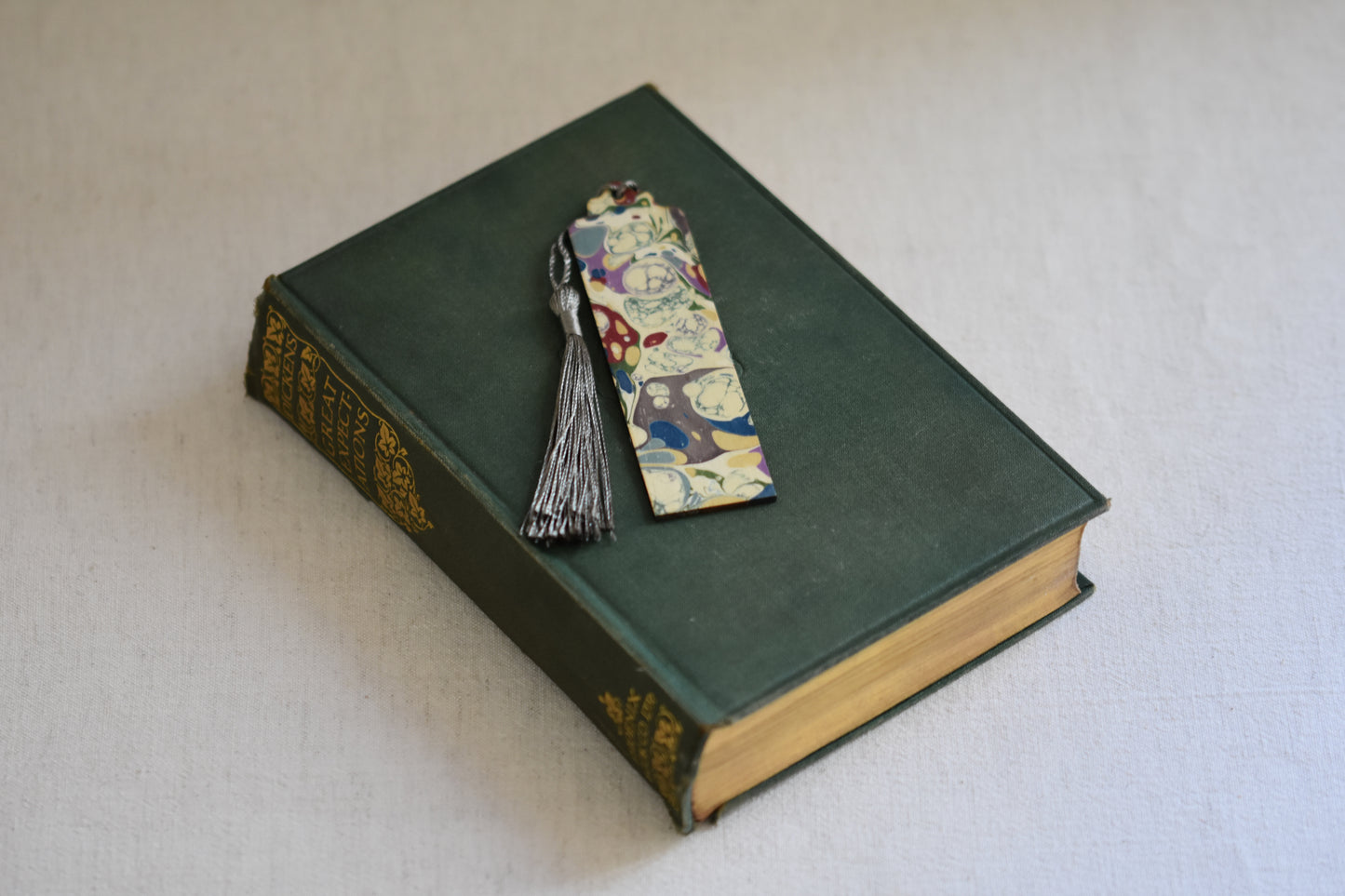 Jemma Lewis colourful marbled wooden bookmark with grey tassel on a vintage green and gold book, with a beige background.
