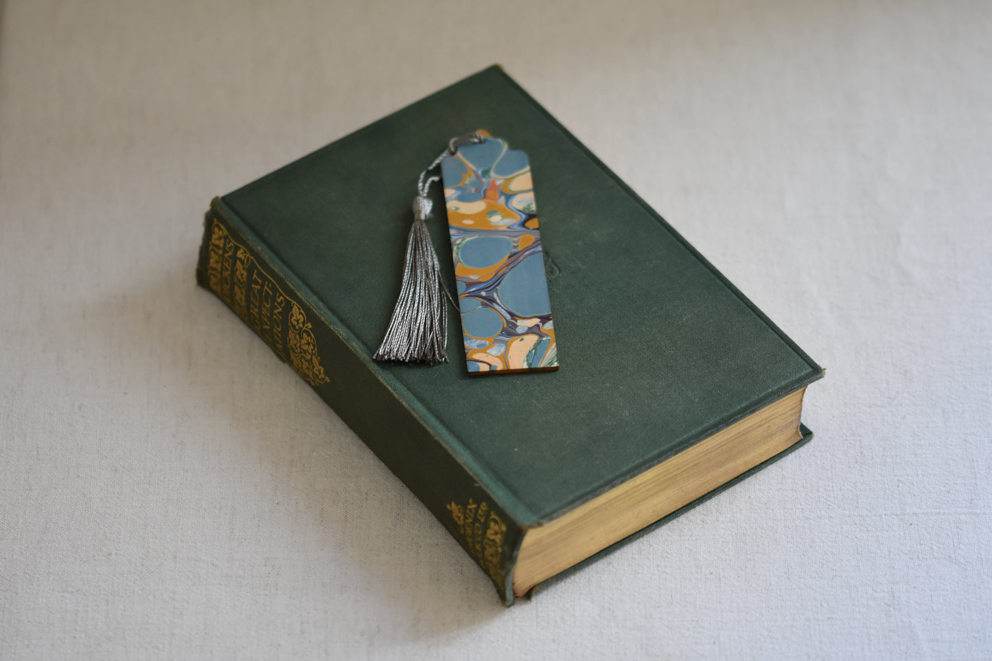 Jemma Lewis colourful marbled wooden bookmark with grey tassel on a vintage green and gold book, with a beige background.
