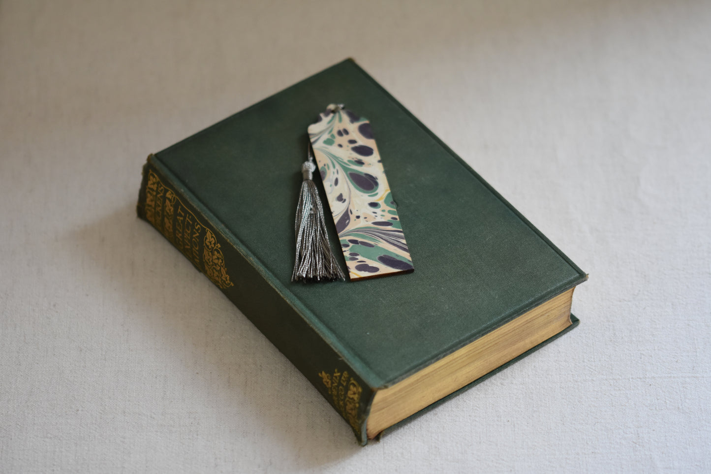 Jemma Lewis colourful marbled wooden bookmark with grey tassel on a vintage green and gold book, with a beige background.