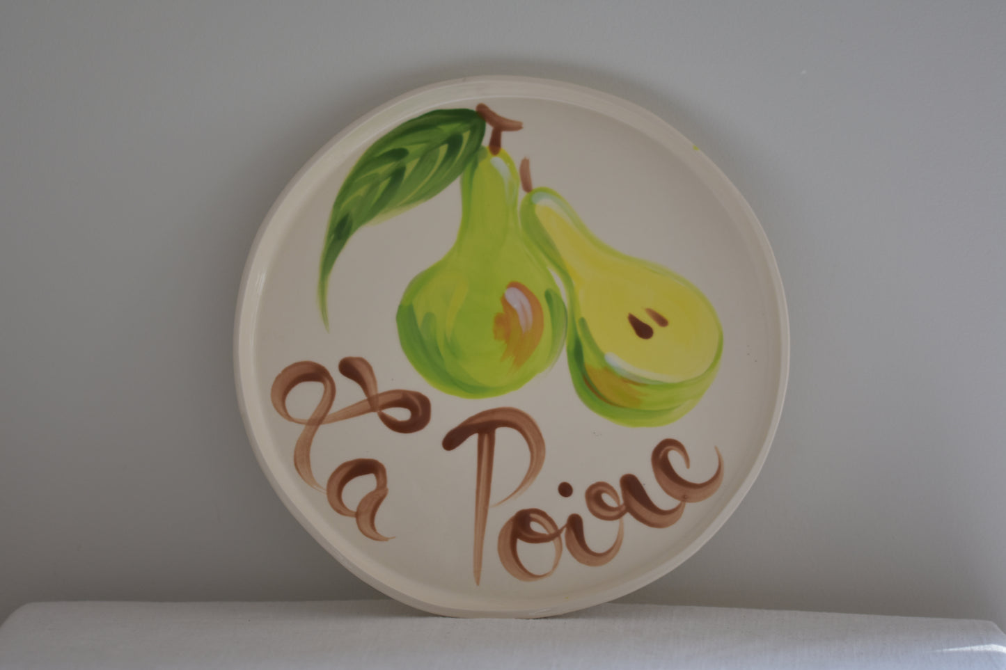 Le Crabe La Poire ceramic plate with painted pear and French writing, on a beige background.