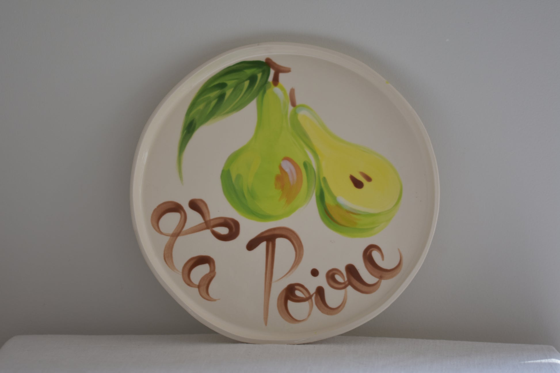 Le Crabe La Poire ceramic plate with painted pear and French writing, on a beige background.