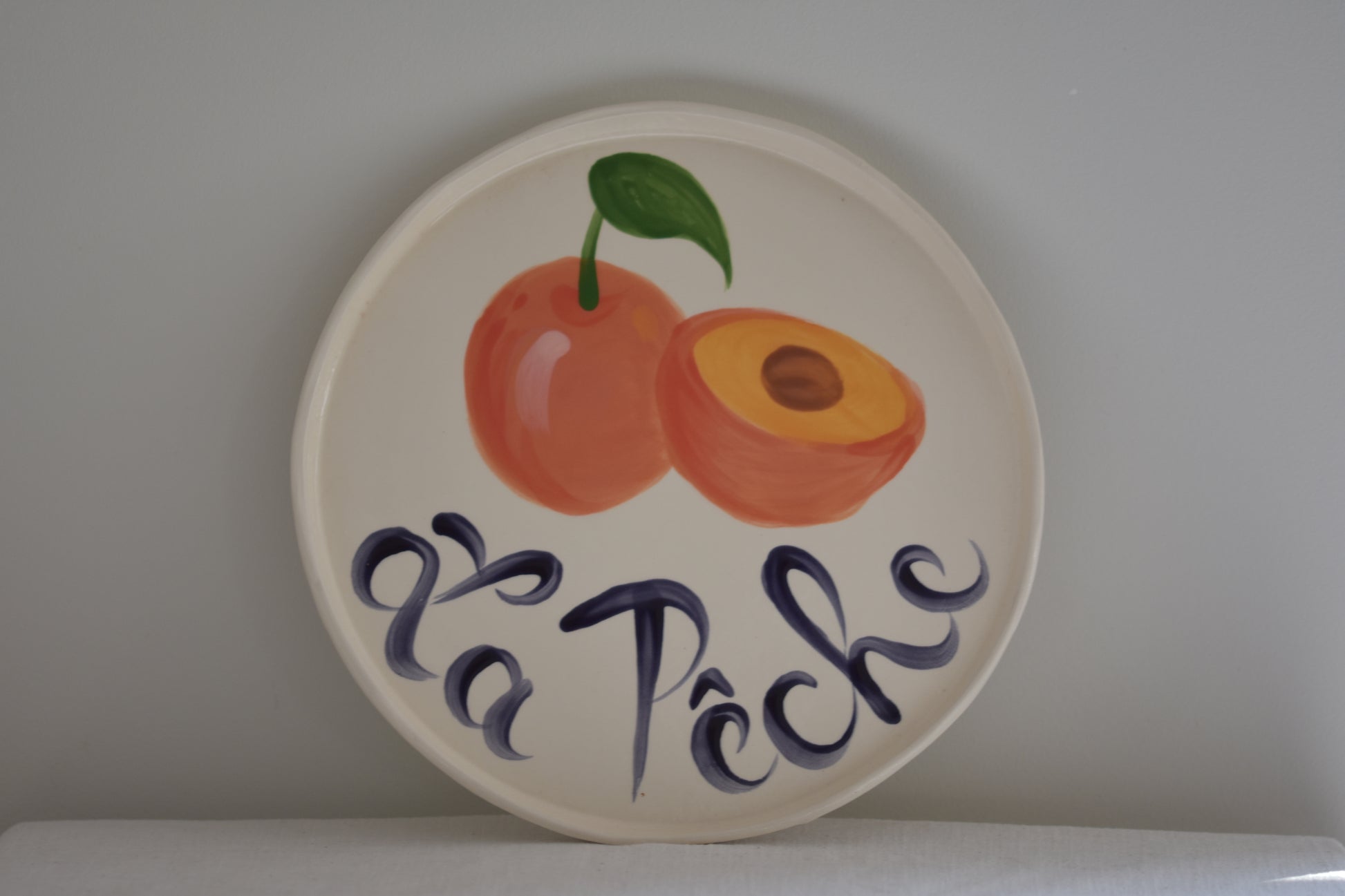 Le Crabe La Peche ceramic plate with painted peach and French writing, on a beige background.