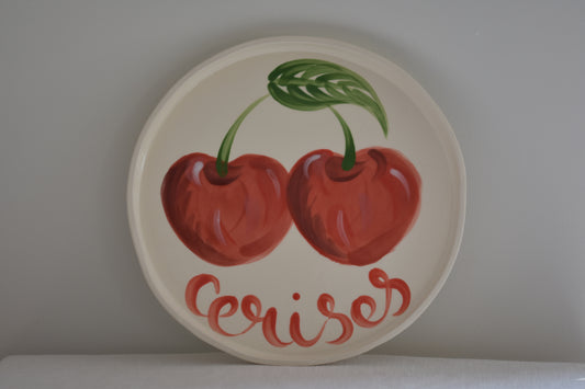 Le Crabe Cerises ceramic plate with painted cherry and French writing, on a beige background.
