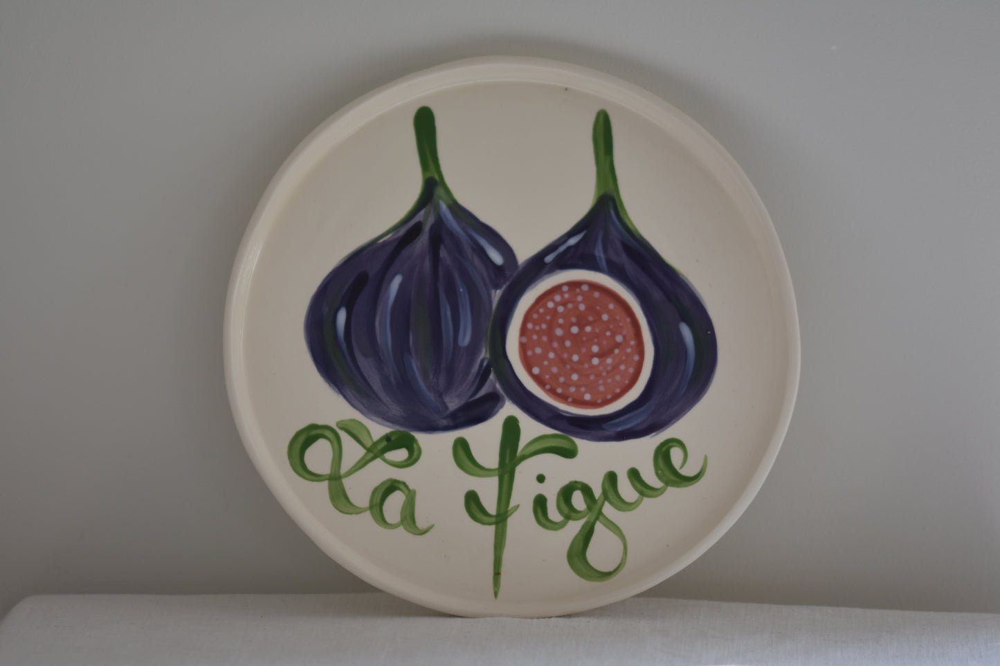 Le Crabe La Figue ceramic plate with painted fig and French writing, on a beige background.