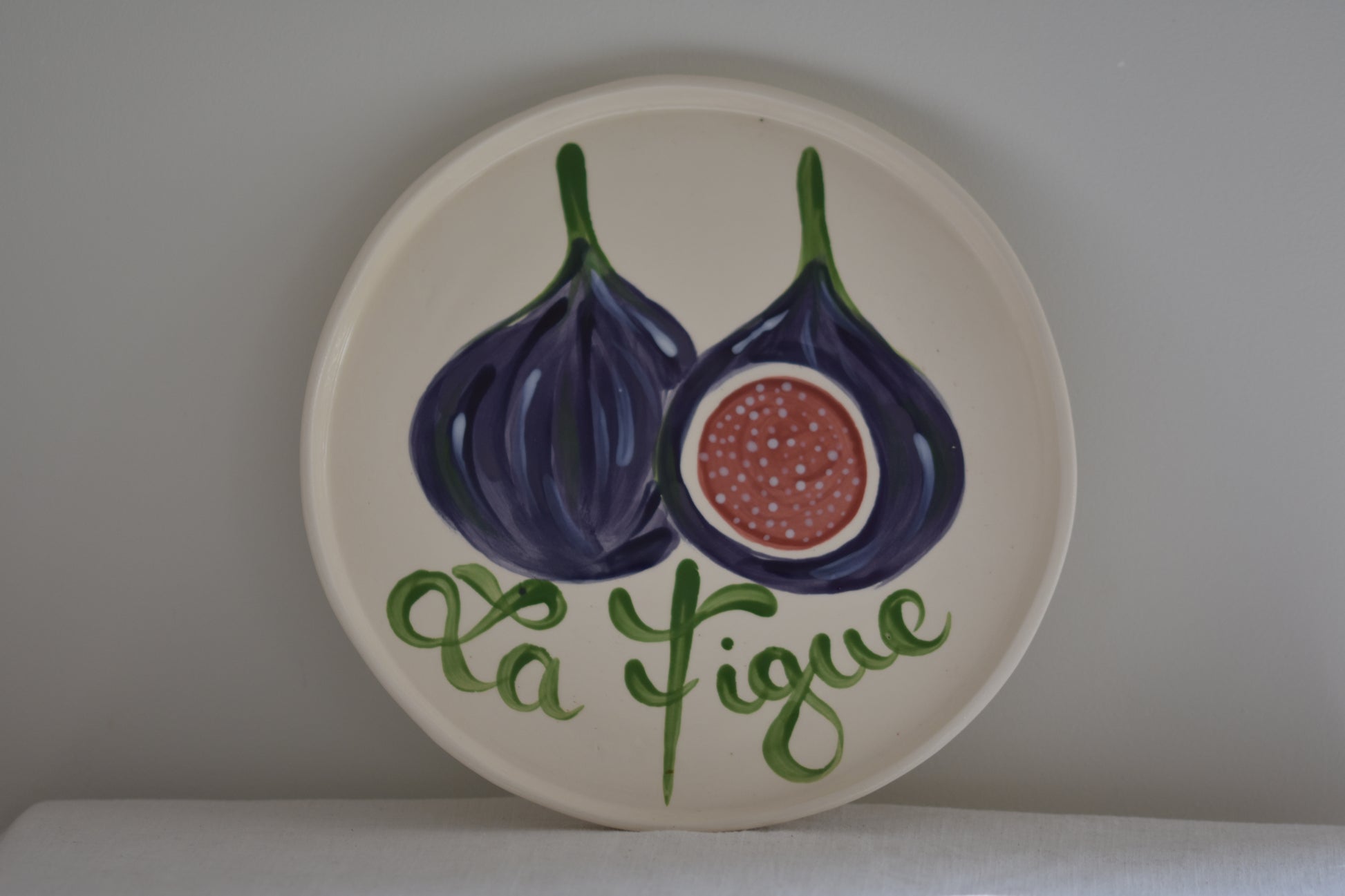 Le Crabe La Figue ceramic plate with painted fig and French writing, on a beige background.