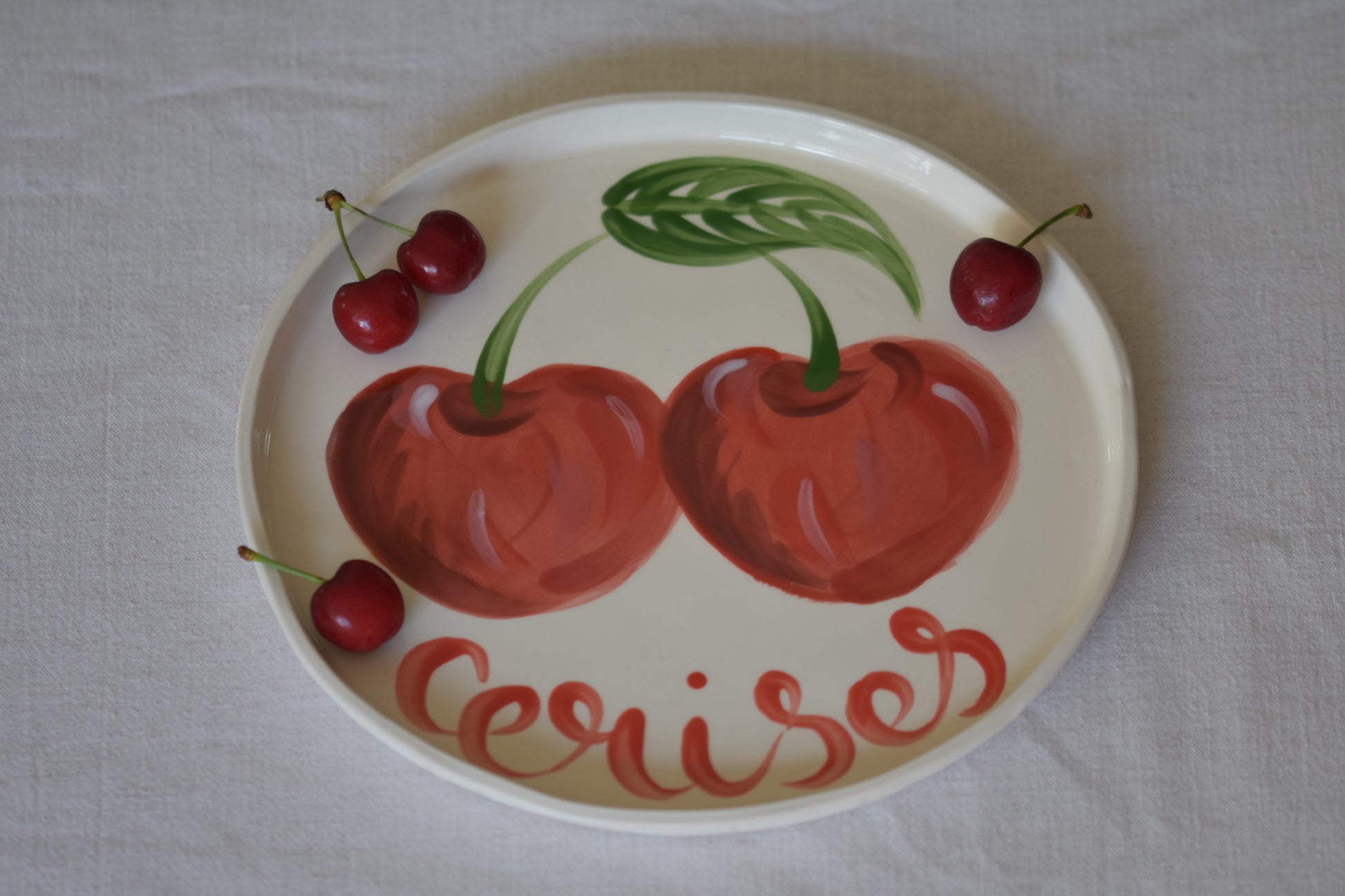 Le Crabe Cerises ceramic plate with painted cherry and French writing, shown with cherries on a beige background.