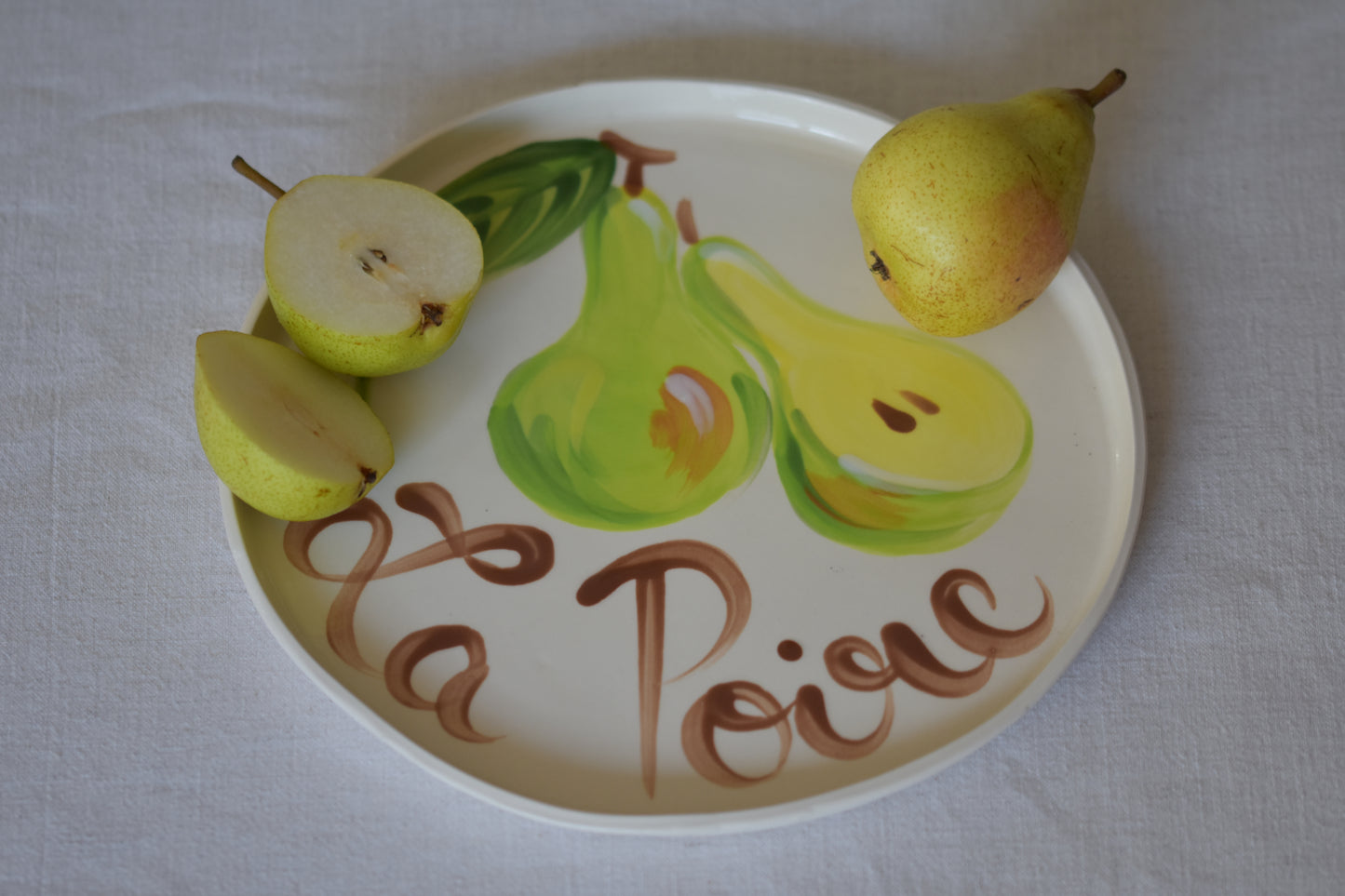 Le Crabe La Poire ceramic plate with painted pear and French writing, shown with pears on a beige background.