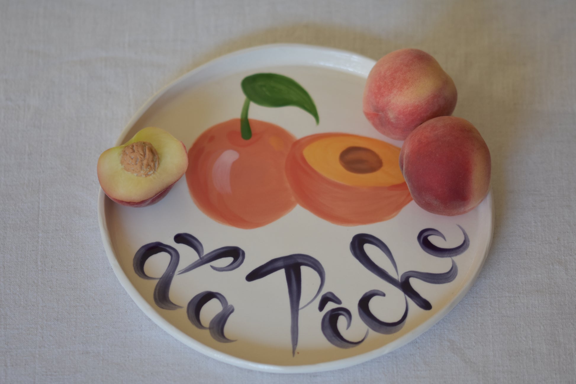 Le Crabe La Peche ceramic plate with painted peach and French writing, shown with peaches on a beige background.
