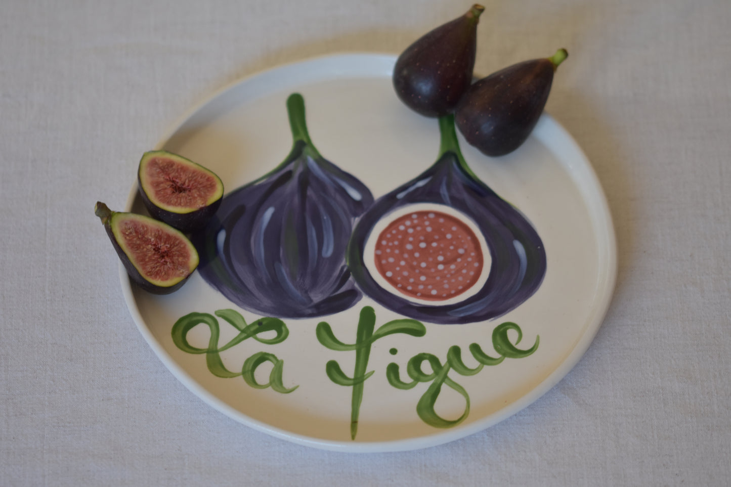 Le Crabe La Figue ceramic plate with painted fig and French writing, shown with figs on a beige background.