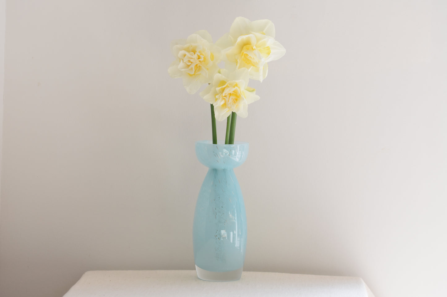 Vintage light blue glass vase containing daffodils, on a beige background.