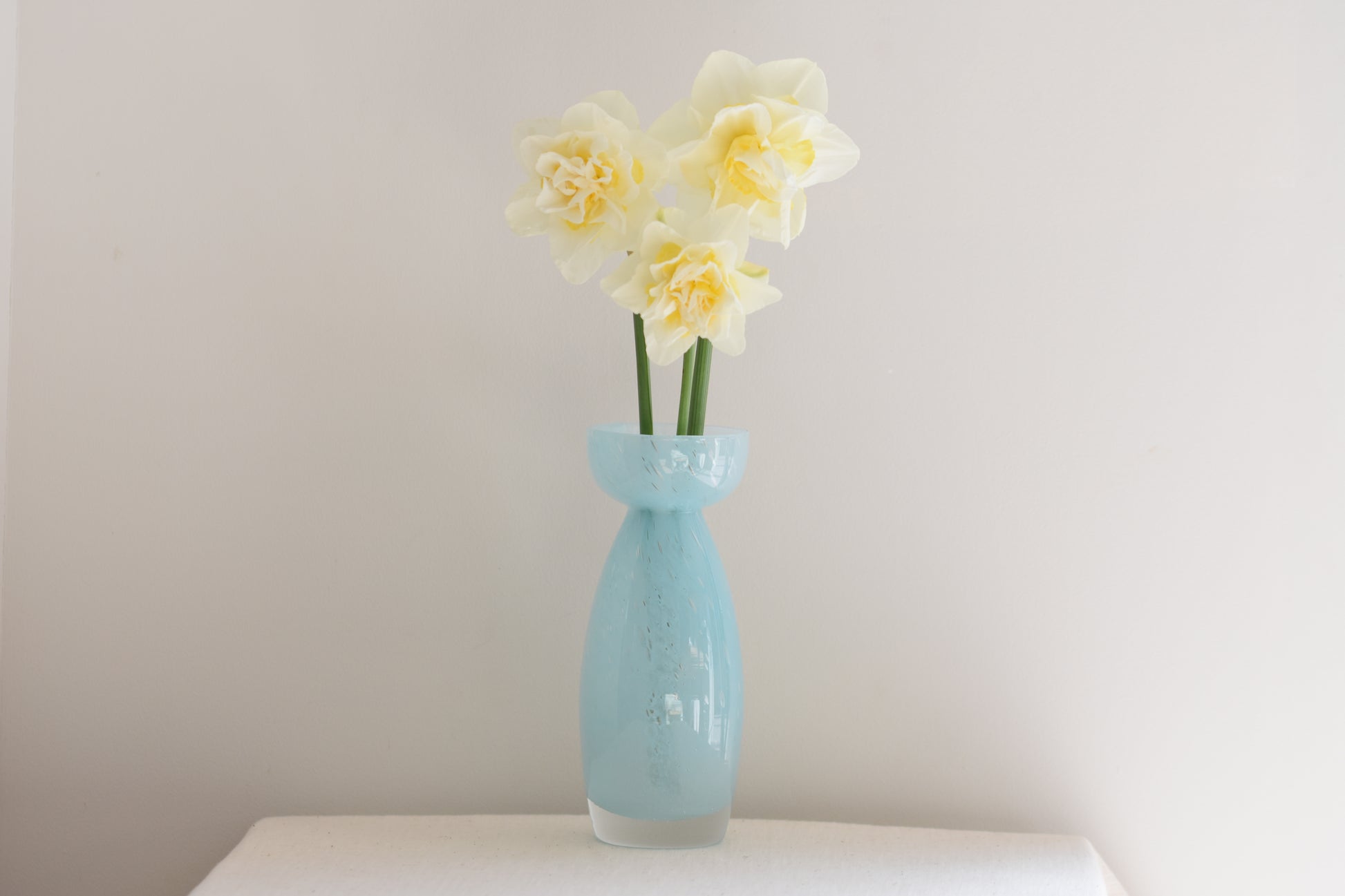 Vintage light blue glass vase containing daffodils, on a beige background.