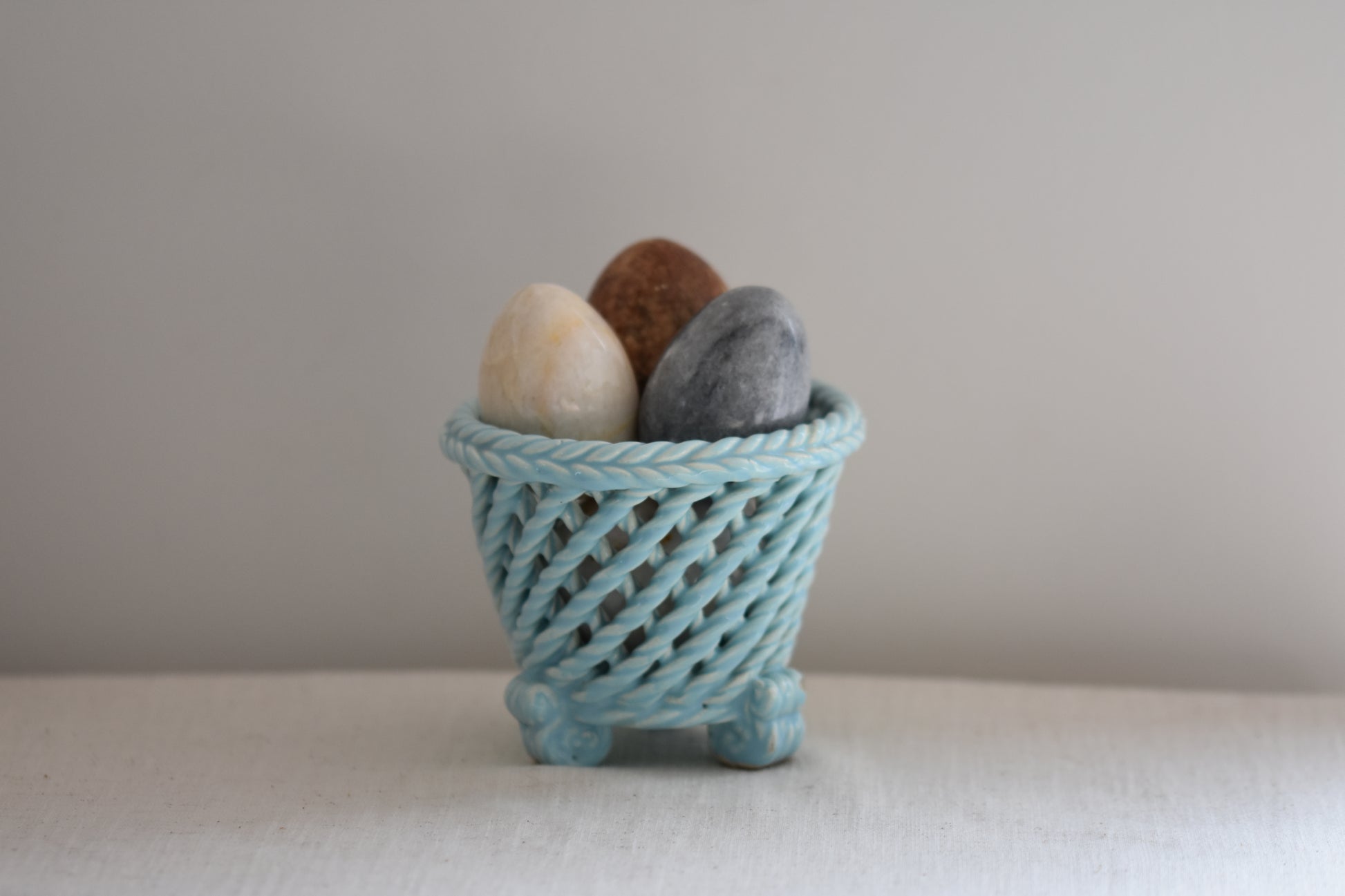 Vintage light blue woven ceramic pot filled with stone eggs, on a beige background.