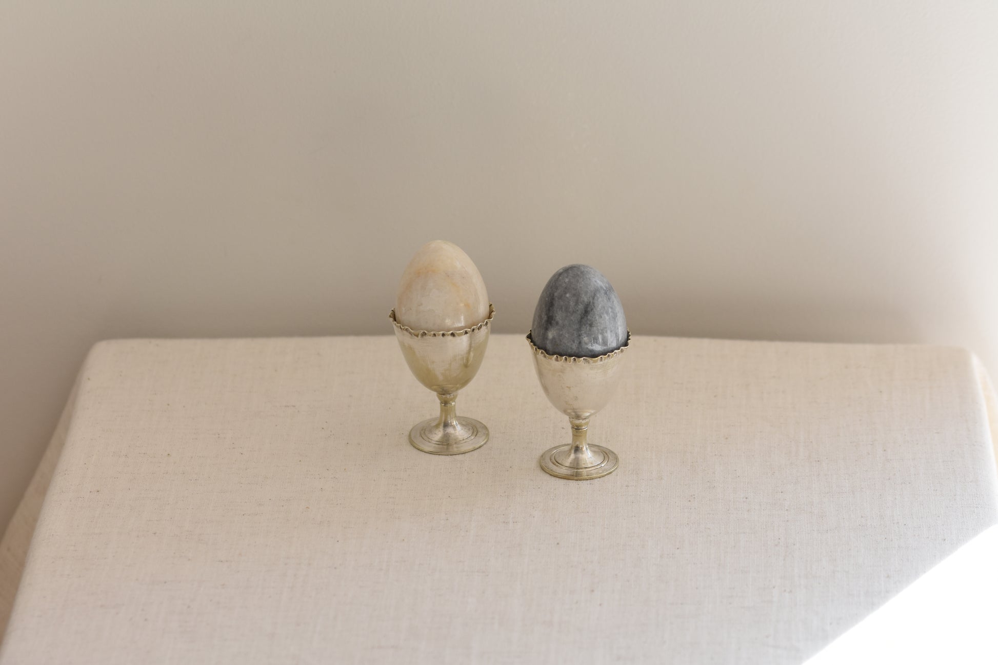Pair of vintage silver plated egg cups with crimped rims holding stone eggs on a beige background.