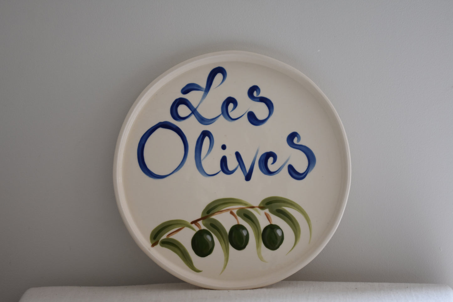 Le Crabe Les Olives ceramic plate with painted olives and French writing, on a beige background.