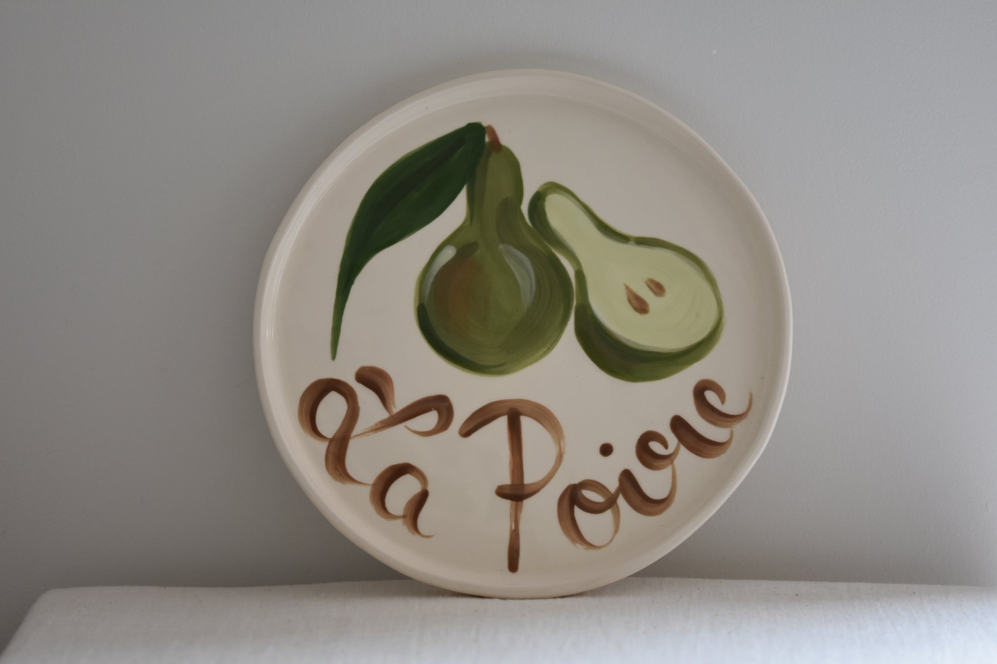 Le Crabe La Poire ceramic plate with painted pear and French writing, on a beige background.
