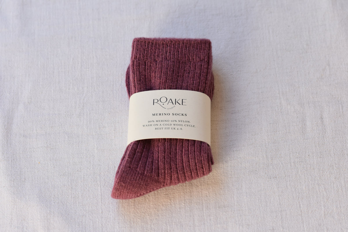 A pair of maroon purple ribbed knit Merino wool socks folded and wrapped with cream paper band reading Roake Studio, on a light fabric background.