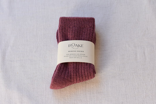 A pair of maroon purple ribbed knit Merino wool socks folded and wrapped with cream paper band reading Roake Studio, on a light fabric background.