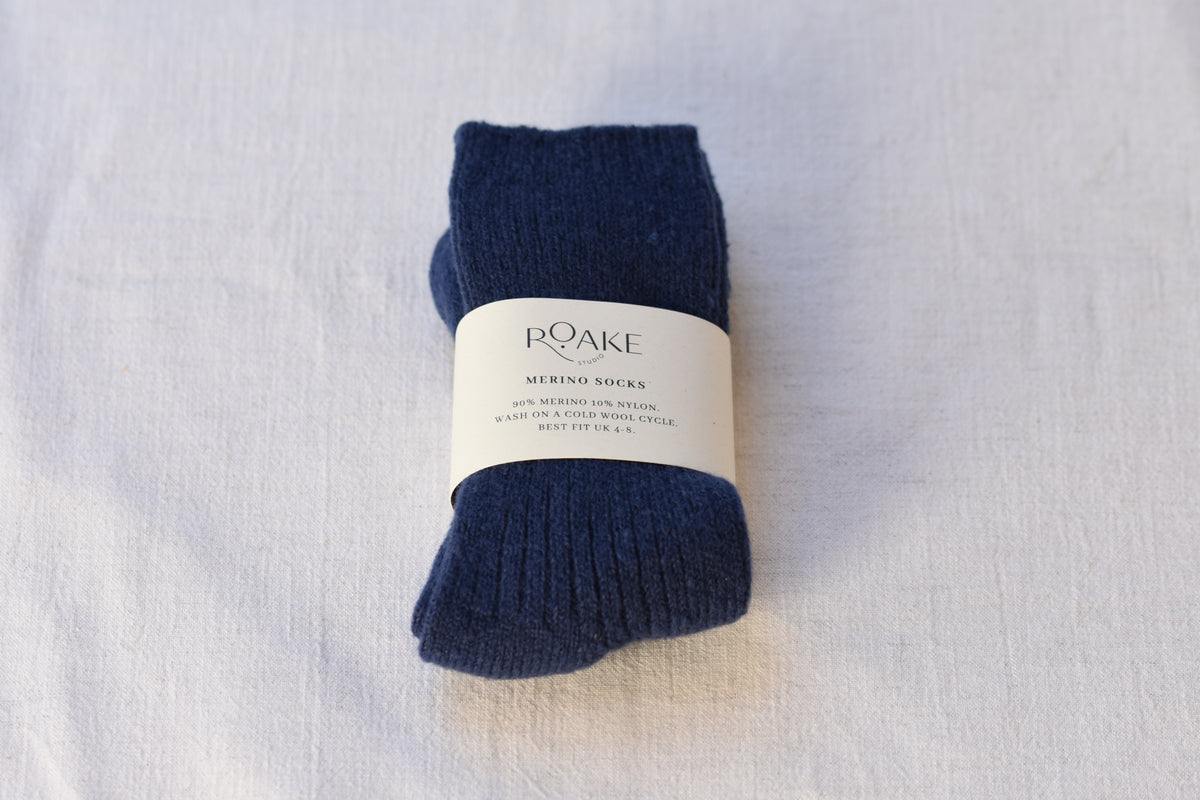 A pair of navy blue ribbed knit Merino wool socks folded and wrapped with cream paper band reading Roake Studio, on a light fabric background.