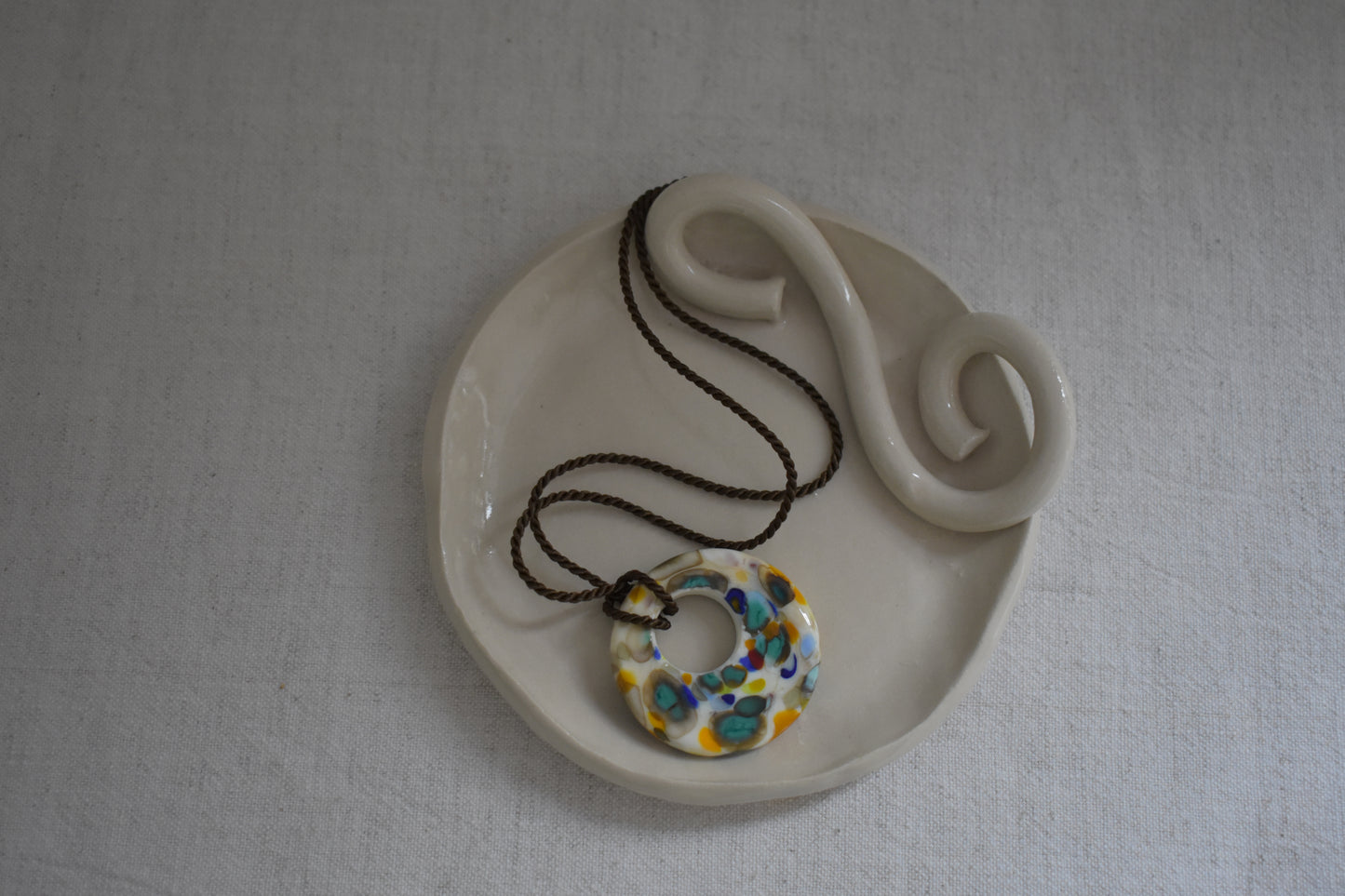 Sandralexandra Zero Waste multicolour glass disc on brown twisted silk cord necklace, show on We Are Clay white ceramic spiral Wee Dish.