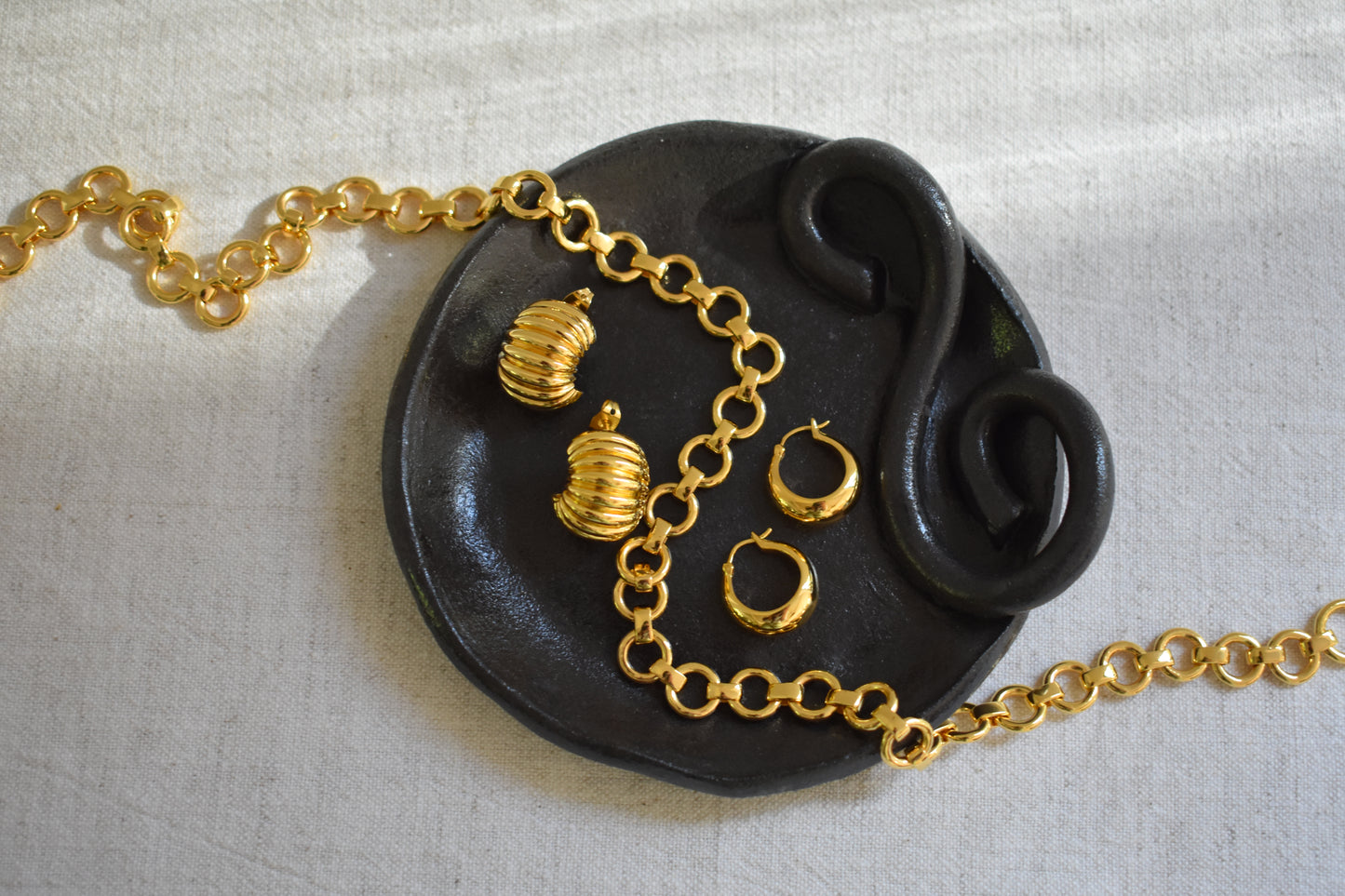 Daphine Baby Bea chain necklace, Mini Oli hoops and Clara hoop earrings in gold on black We Are Clay ceramic spiral Wee dish.