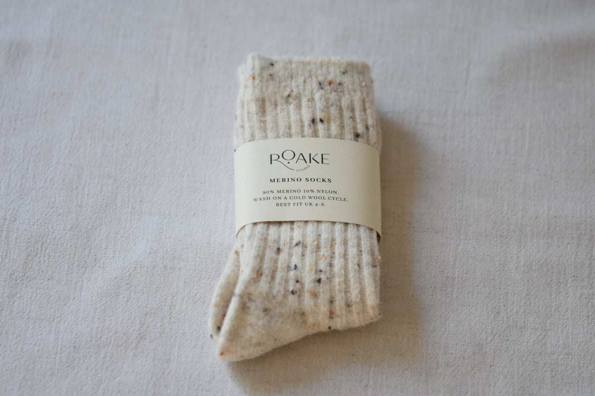 A pair of cream fleck ribbed knit Merino wool socks folded and wrapped with cream paper band reading Roake Studio, on a light fabric background.