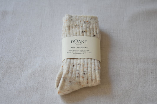 A pair of cream fleck ribbed knit Merino wool socks folded and wrapped with cream paper band reading Roake Studio, on a light fabric background.